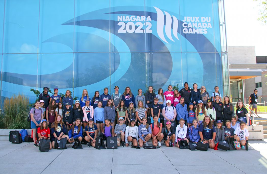 About 50 young women sit in front of a large blue backdrop that says Niagara 2022 Canada Games.