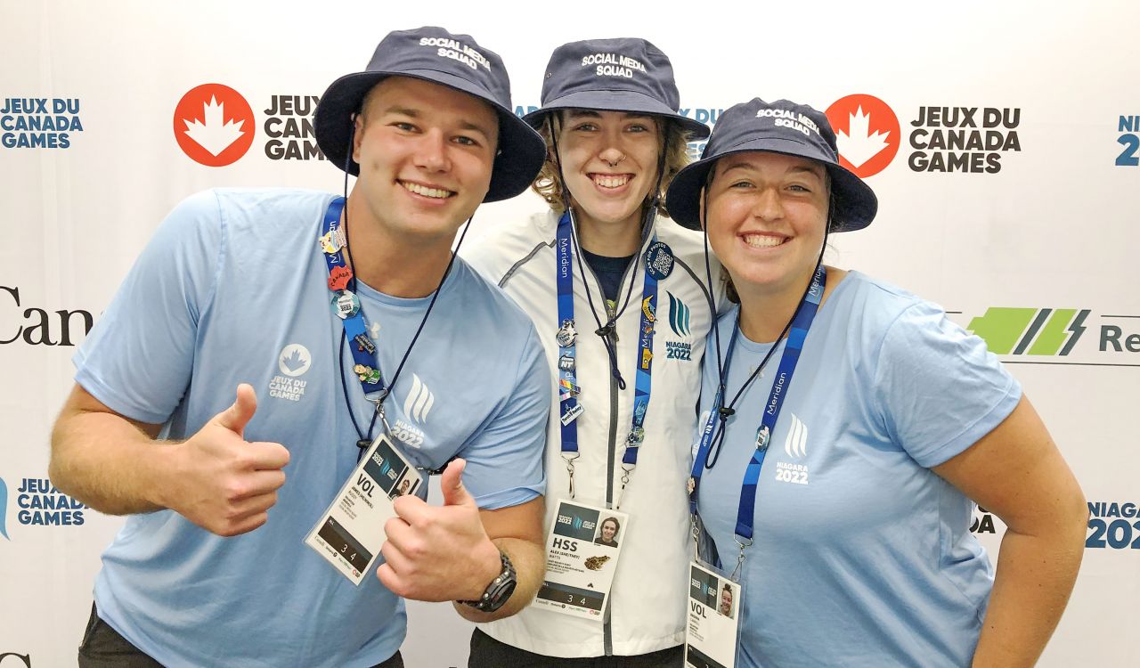 Three students in bucket hats and lanyards stand in front of a promotional backdrop featuring logos for the Province of Ontario, Niagara Region and the Niagara 2022 Canada Summer Games.