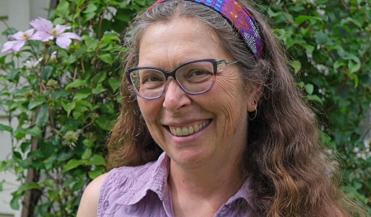 Miriam Richards headshot, standing in front of green foliage wall wearing purple shirt with matching patterned headband and dark frame glasses.