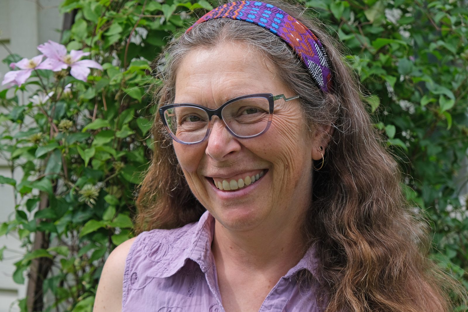 Miriam Richards headshot, standing in front of green foliage wall wearing purple shirt with matching patterned headband and dark frame glasses.