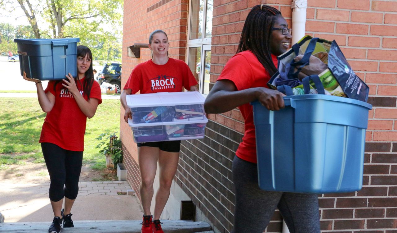 Three women carrying large bins walk past a brick building.