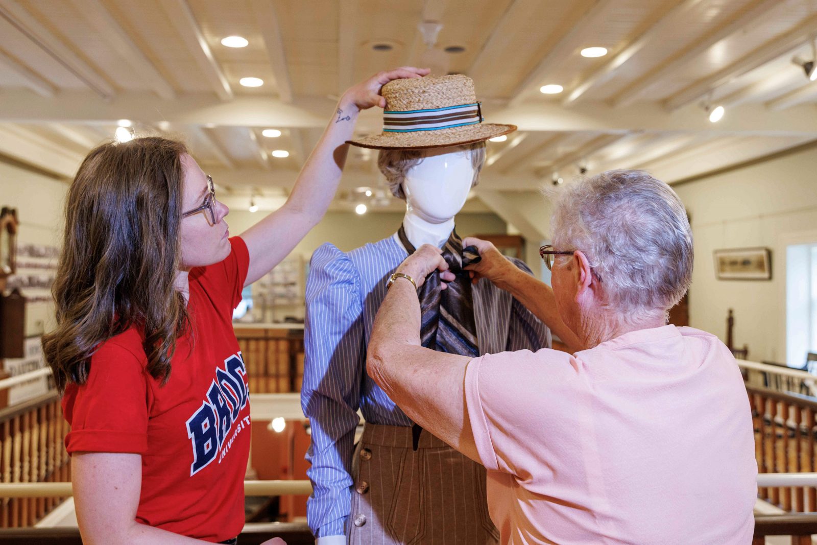 Two women put old-fashioned clothing and a hat on a mannequin.
