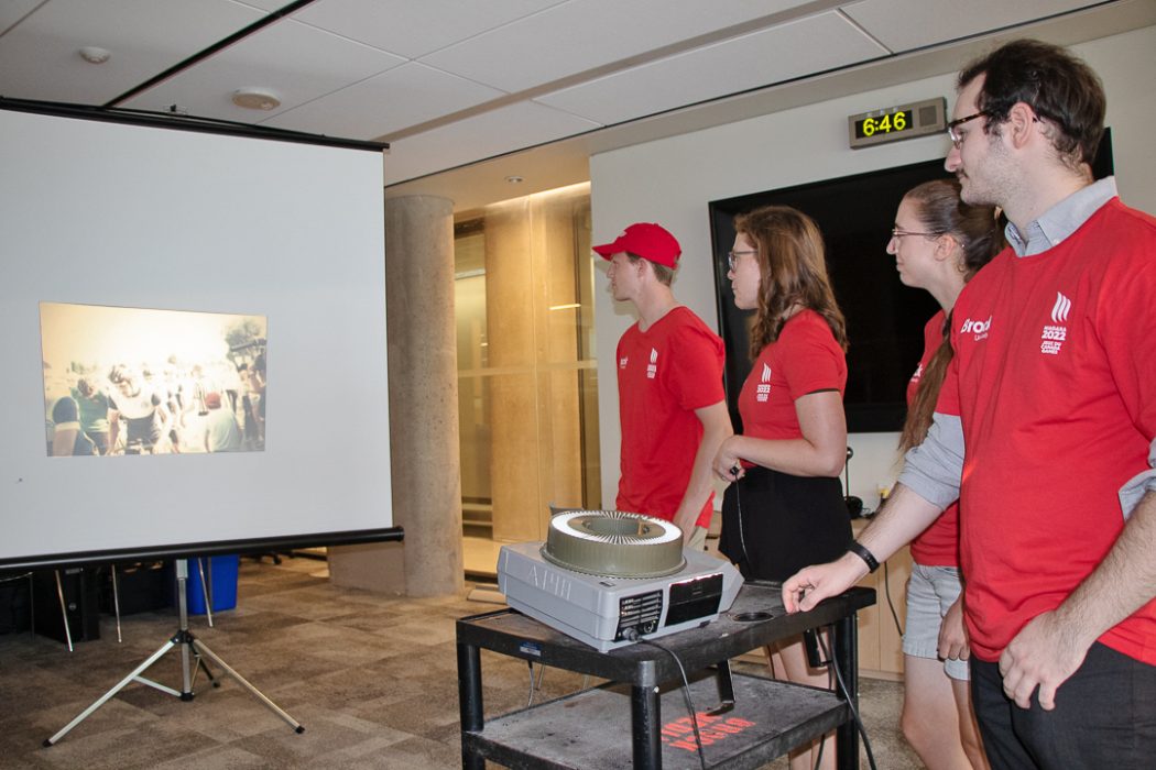 Four people stand looking at a projector screen that has an old image of cyclists on it.