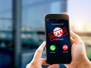 Hands hold a mobile phone. The screen shows an incoming call from an unknown number. The options are to accept or decline. A warning message written in red, large text that reads “SCAM!” is over top of an unknown caller avatar.
