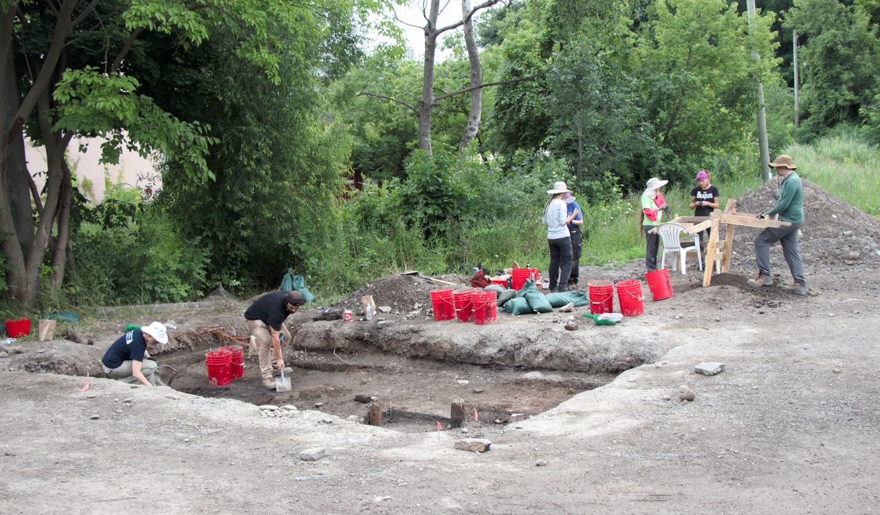 A group of people stand on an archaeological dig site that has a large rectangular area dug out of the ground. A number of red buckets sit on the raised soil.