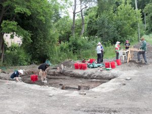 A group of people stand on an archaeological dig site that has a large rectangular area dug out of the ground. A number of red buckets sit on the raised soil.