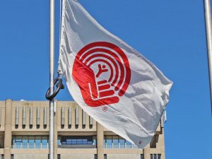 The United Way flag flies in front of Brock University’s Schmon Tower. The flag is white with the red United Way logo, which looks like a hand holding a stick figure while a rainbow arches over the figure. Only the top of the Schmon Tower building is visible in the background. A clear blue sky takes up the rest of the frame.