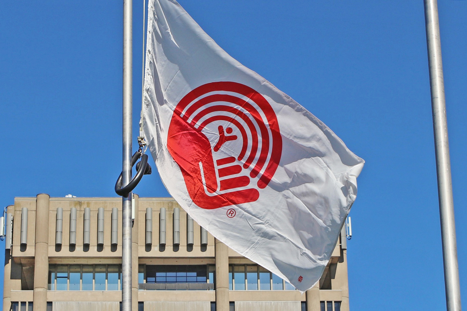 The United Way flag flies in front of 全球电竞直播 ’s Schmon Tower. The flag is white with the red United Way logo, which looks like a hand holding a stick figure while a rainbow arches over the figure. Only the top of the Schmon Tower building is visible in the background. A clear blue sky takes up the rest of the frame.