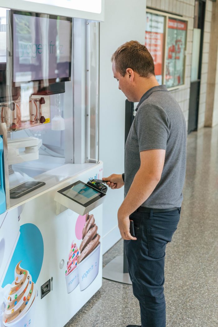 A man taps a card on a pinpad in front of a robotic frozen yogurt machine with a large glass window and robotic arms behind the glass.