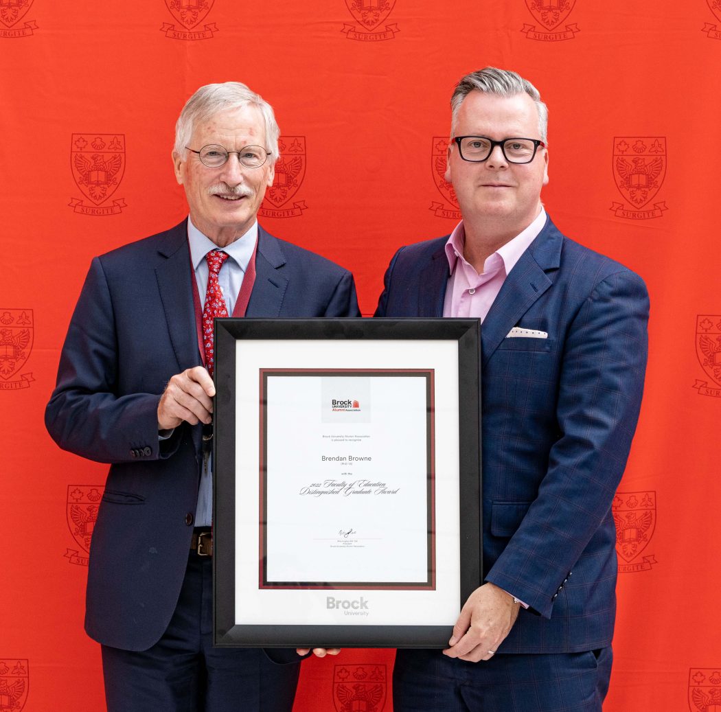 Two men in suits stand together holding a framed certificate.