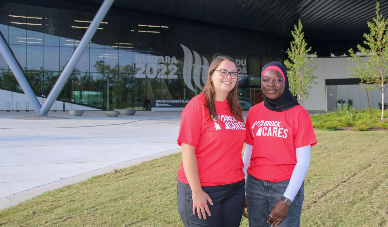 Two women in red T-shirts that say ‘Brock Cares’ on them stand in front of plaza with a glass wall in the background.