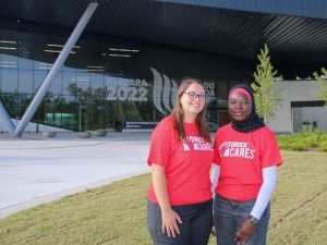 Two women in red T-shirts that say ‘Brock Cares’ on them stand in front of plaza with a glass wall in the background.