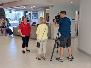 A woman in a red shirt speaks to a reporter and recording camera in the hallway of a residence building