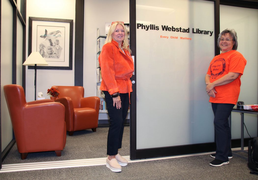 Two women in orange shirts stand in front of a sign that says ‘Phyllis Webstad Library.’
