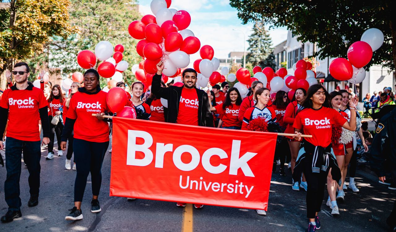 A group of people in red shirts walk together with balloons and a sign that says 'Brock University.'