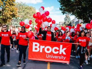 A group of people in red shirts walk together with balloons and a sign that says 'Brock University.'