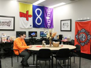 A man in an orange shirt sits writing at a table in front of a yellow, white and red flag, a blue and white flag, and a purple and white flag, as well as an orange flag that says, ‘Every child matters.’