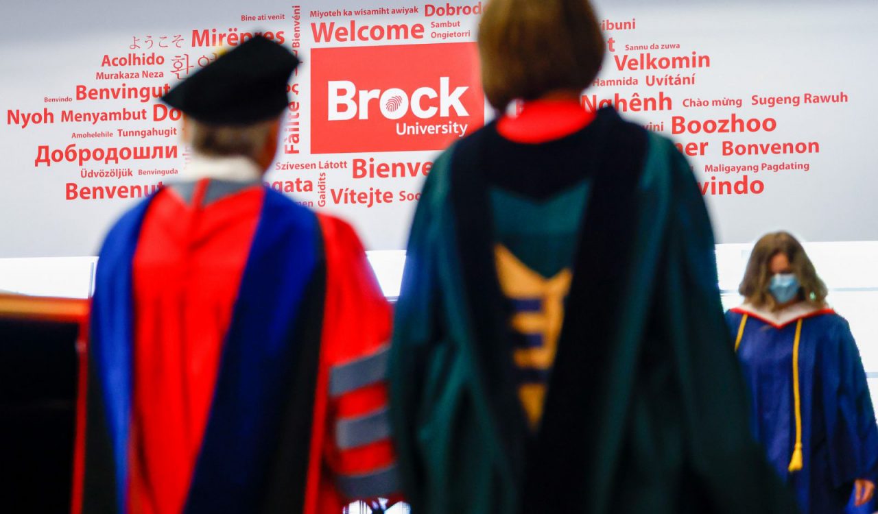 Blurred figures wearing red and black Convocation regalia face a wall with the word ‘welcome’ written in many languages.