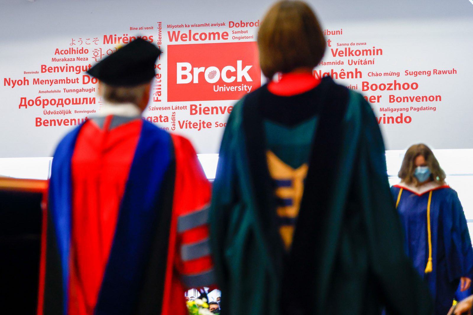 Blurred figures wearing red and black Convocation regalia face a wall with the word ‘welcome’ written in many languages.