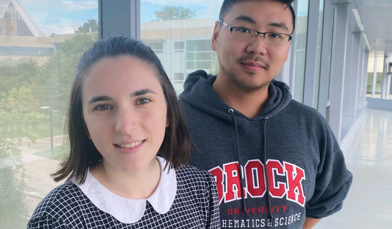 Two students, Melanie Denomme and Noah Xiao, pose in a long corridor with glass windows overlooking Brock University campus.