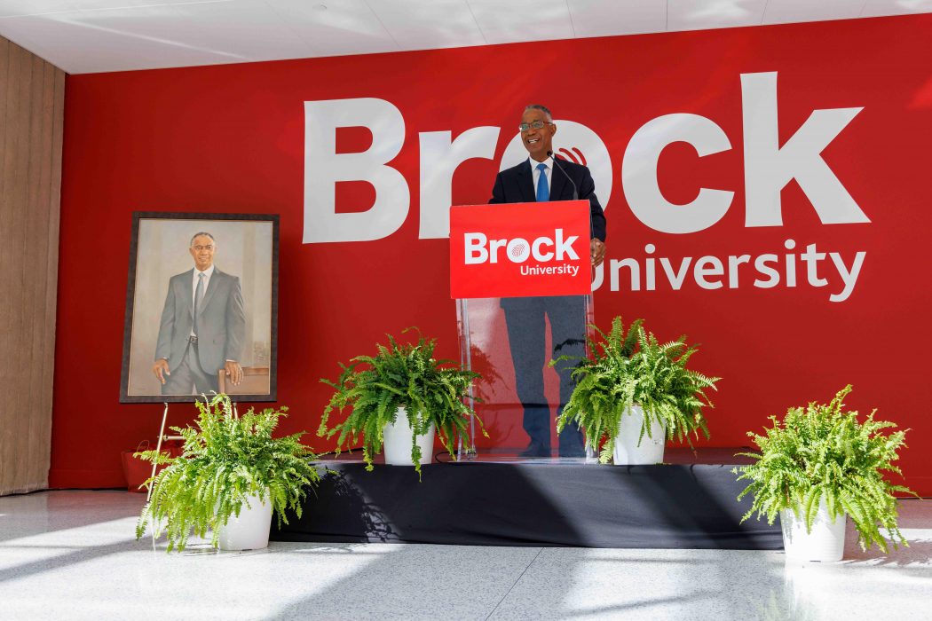 A man stands at a podium speaking with a large Brock University sign in the background and a painting of the man on the left.