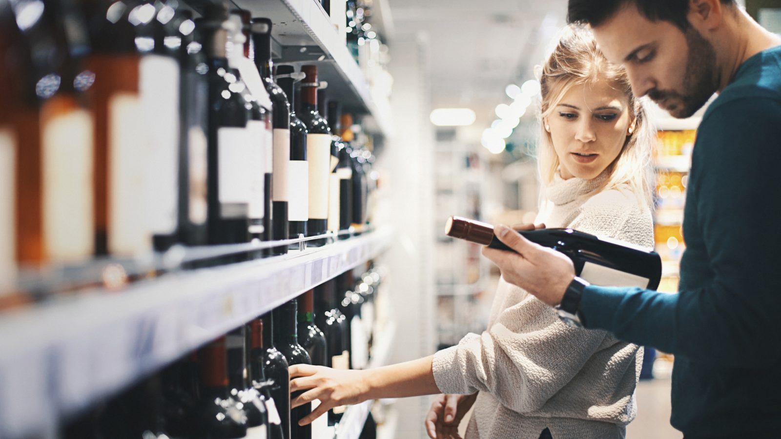 A man and woman stand in front of store shelves filled with wine bottles. The man is holding a bottle and looking at the label.