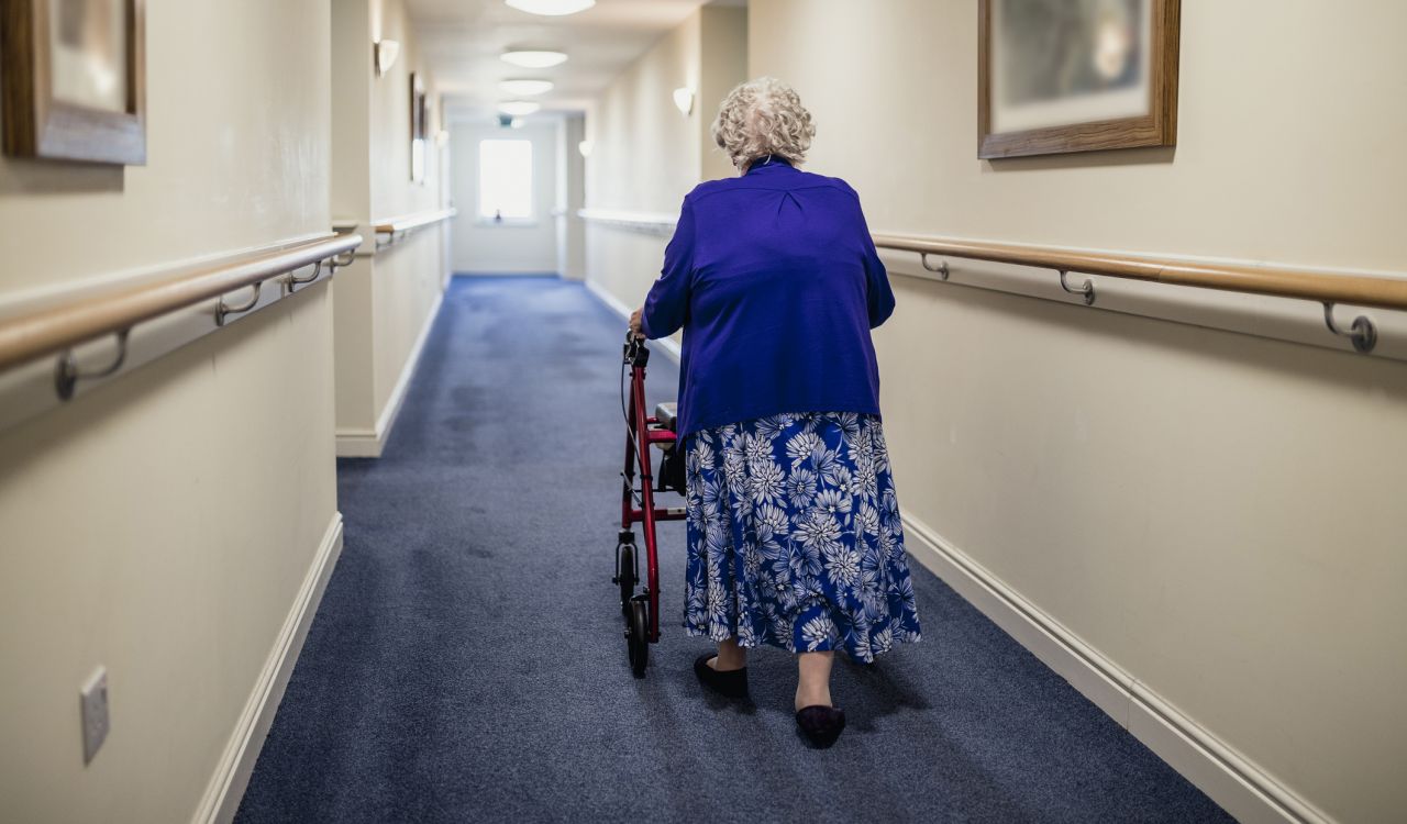 An older adult woman wearing a blue jacket and blue patterned skirt and using a red walker walks down a blue-carpeted hall in a long-term care facility; view from behind.