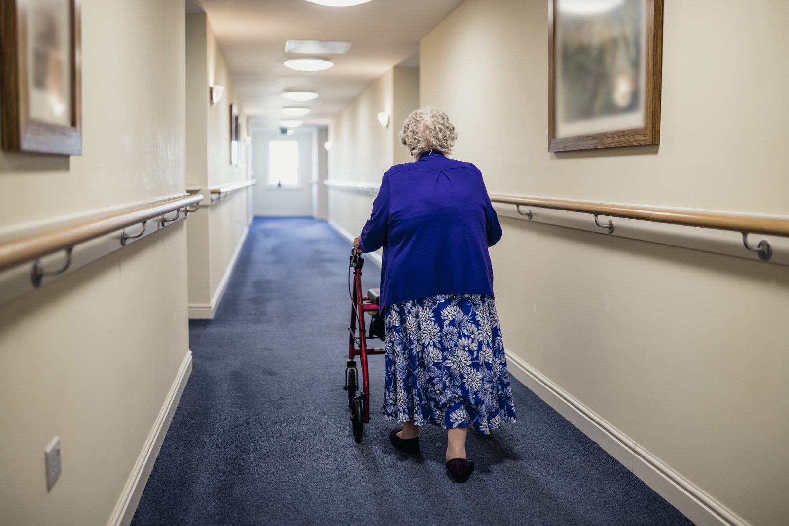 An older adult woman wearing a blue jacket and blue patterned skirt and using a red walker walks down a blue-carpeted hall in a long-term care facility; view from behind.