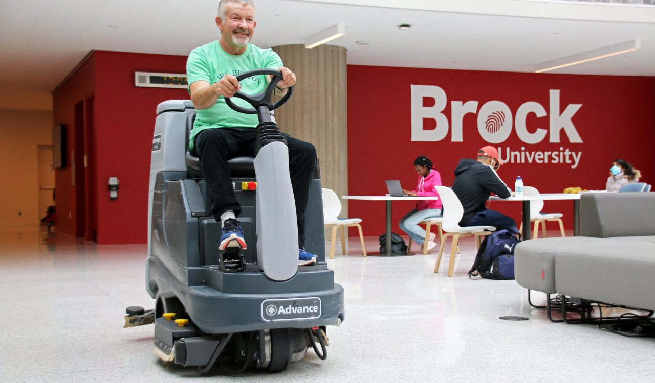 A person sits atop a ride-on automatic floor scrubber to clean the floors in a large, naturally lit building. In the background are several people sitting at tables and chairs in front of a large red wall with “Brock University” written in white.