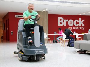 A person sits atop a ride-on automatic floor scrubber to clean the floors in a large, naturally lit building. In the background are several people sitting at tables and chairs in front of a large red wall with “Brock University” written in white.