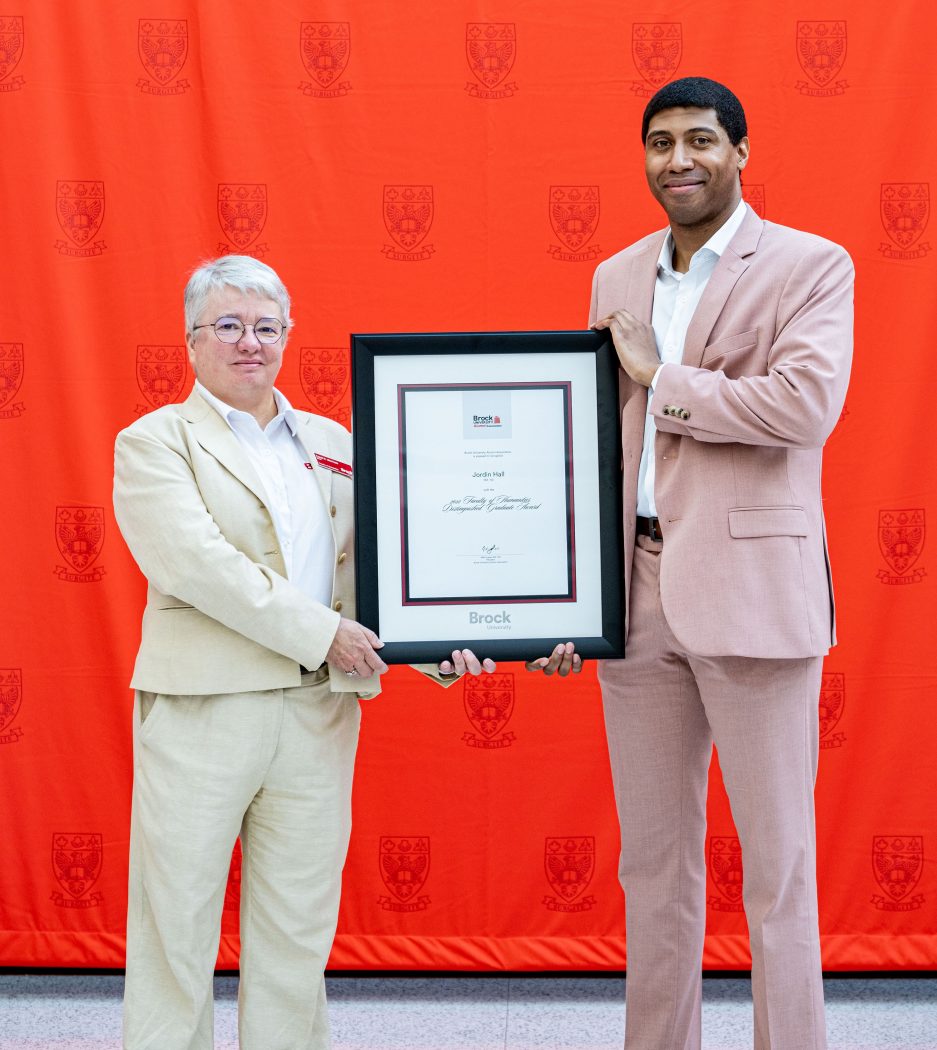 A woman on the left presenting a framed certificate to a male on the right pictured in front of a red backdrop.