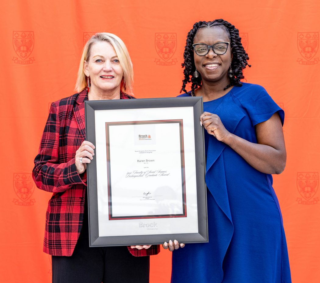 Two women hold an award between them while standing in front of a red backdrop.