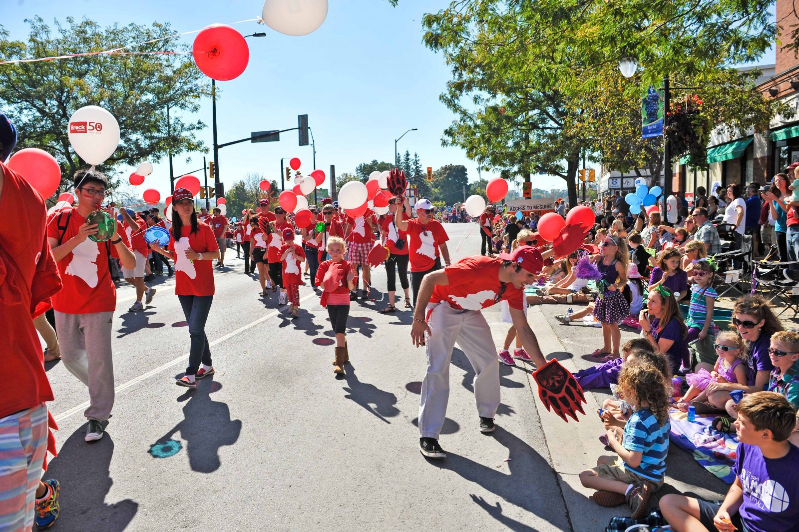 A crowd of people in red shirts carrying red balloons walks along a parade route lined with people.