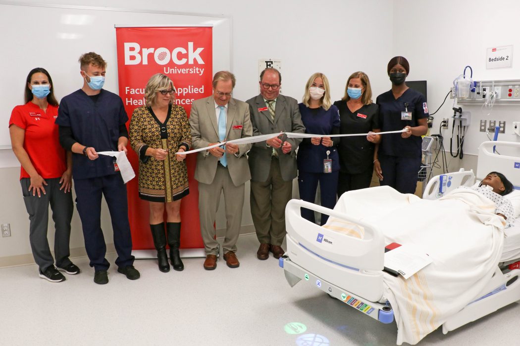 A group of eight people stood in a row while the man in the middle cut off a piece of gauze.  The group stands next to a patient simulator in a lab that looks and feels like a hospital room.