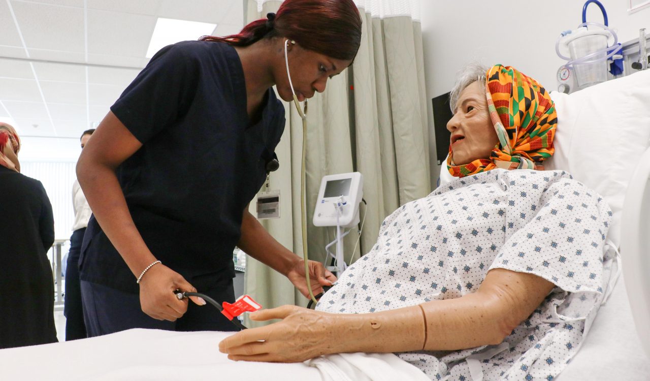 A nurse examines a patient simulator in a nursing lab designed to look like a hospital room.