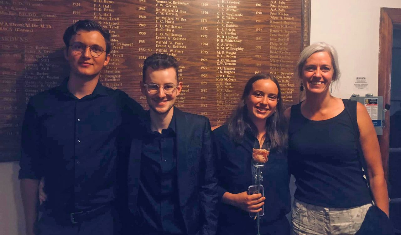 An image of a group of four individuals: two men on the left and two women on the right, all wearing black semi-formal wear. There is a wooden wall behind them with names of recognized individuals and years.