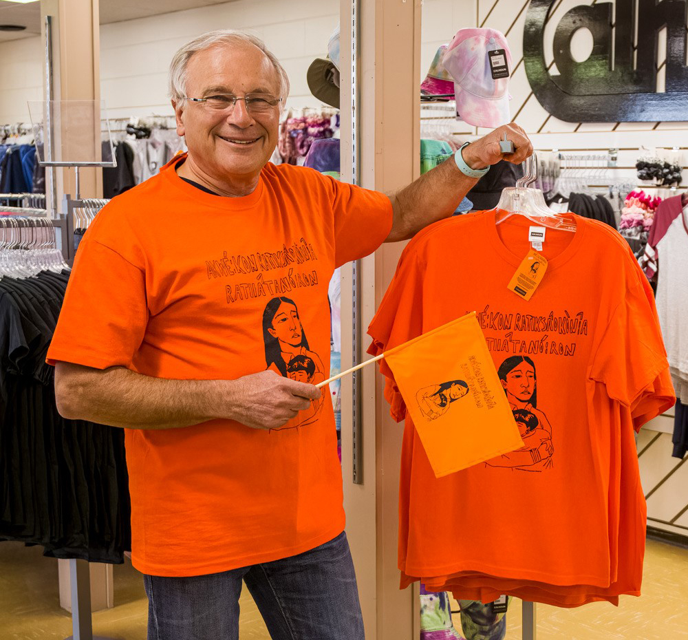 A man in an orange shirt stands with a display of orange shirts and holds an orange flag.