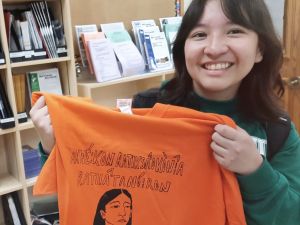 A smiling woman holds an orange T-shirt with words written in the Mohawk language.
