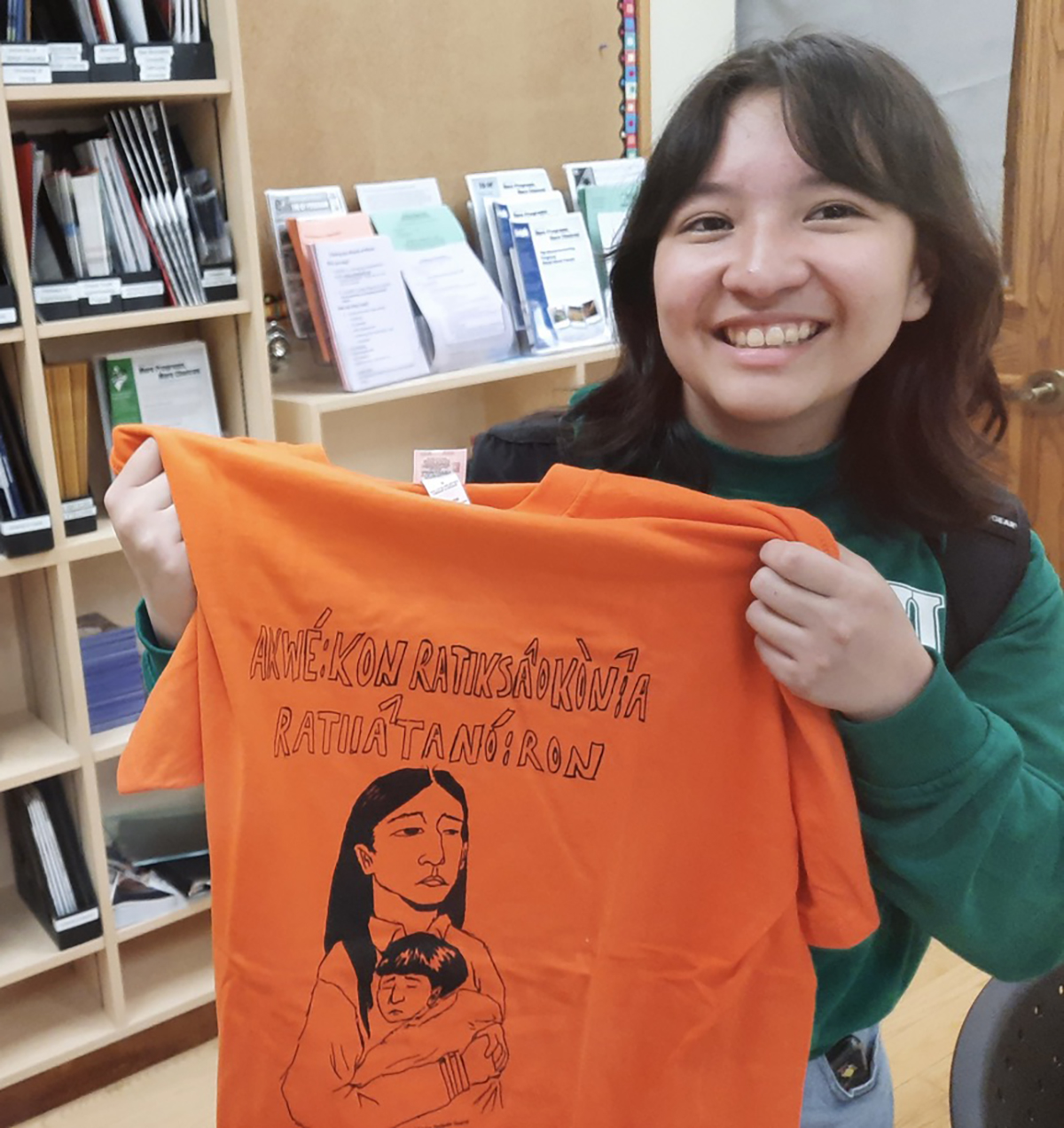 A smiling woman holds an orange T-shirt with words written in the Mohawk language.
