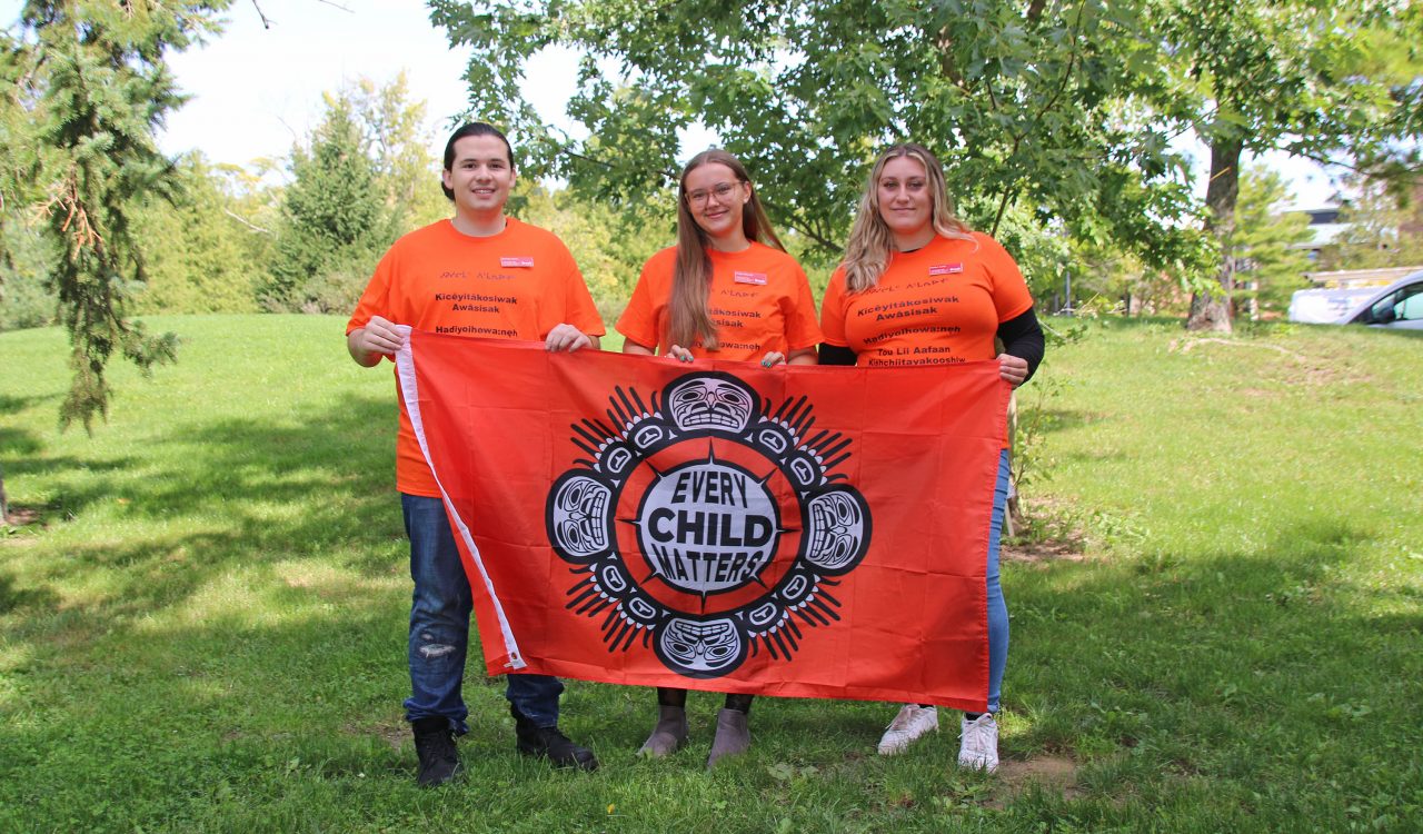 A man and two women stand in an outdoor setting wearing orange shirts and holding a flag that says ‘Every Child Matters.’