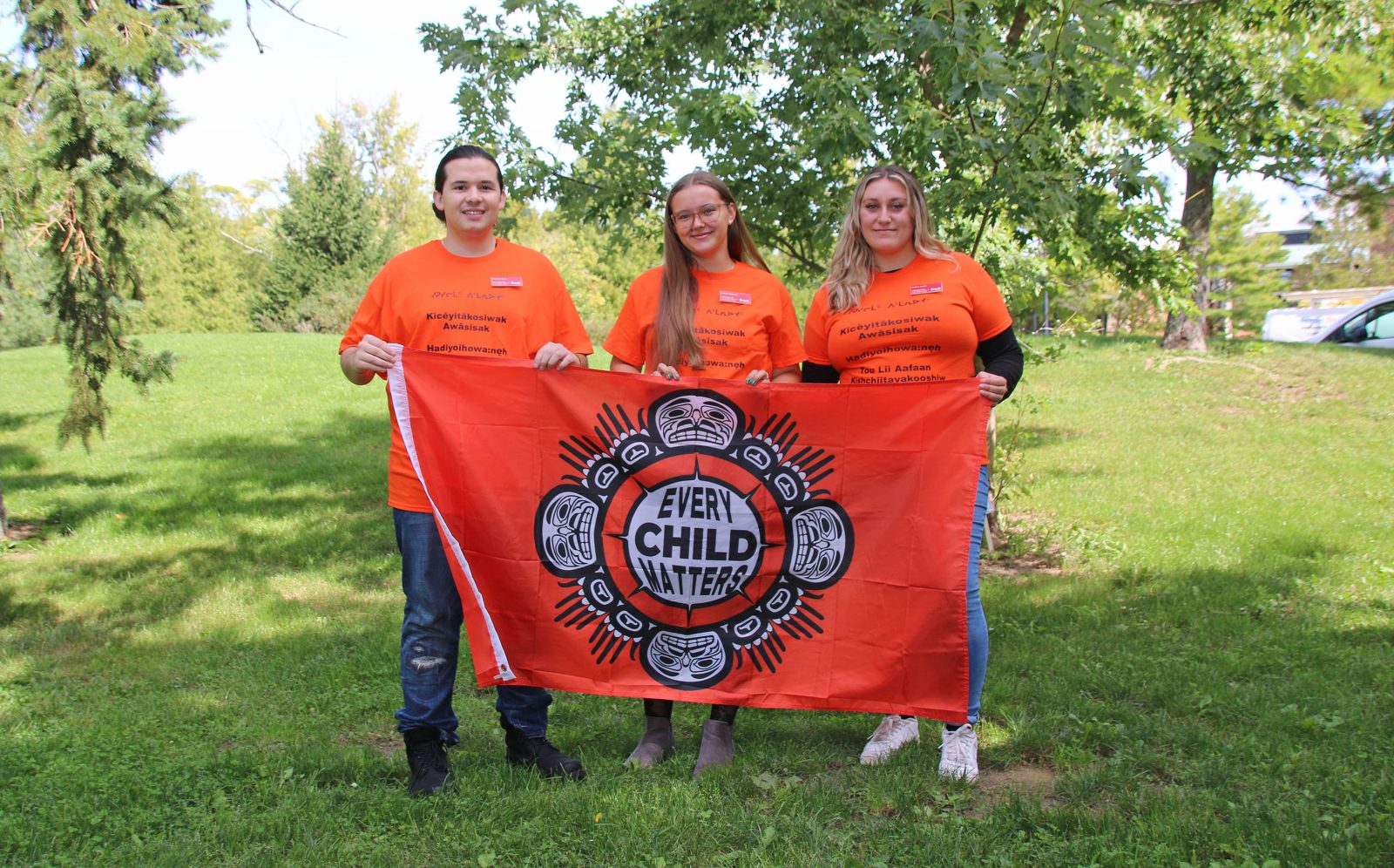 A man and two women stand in an outdoor setting wearing orange shirts and holding a flag that says ‘Every Child Matters.’