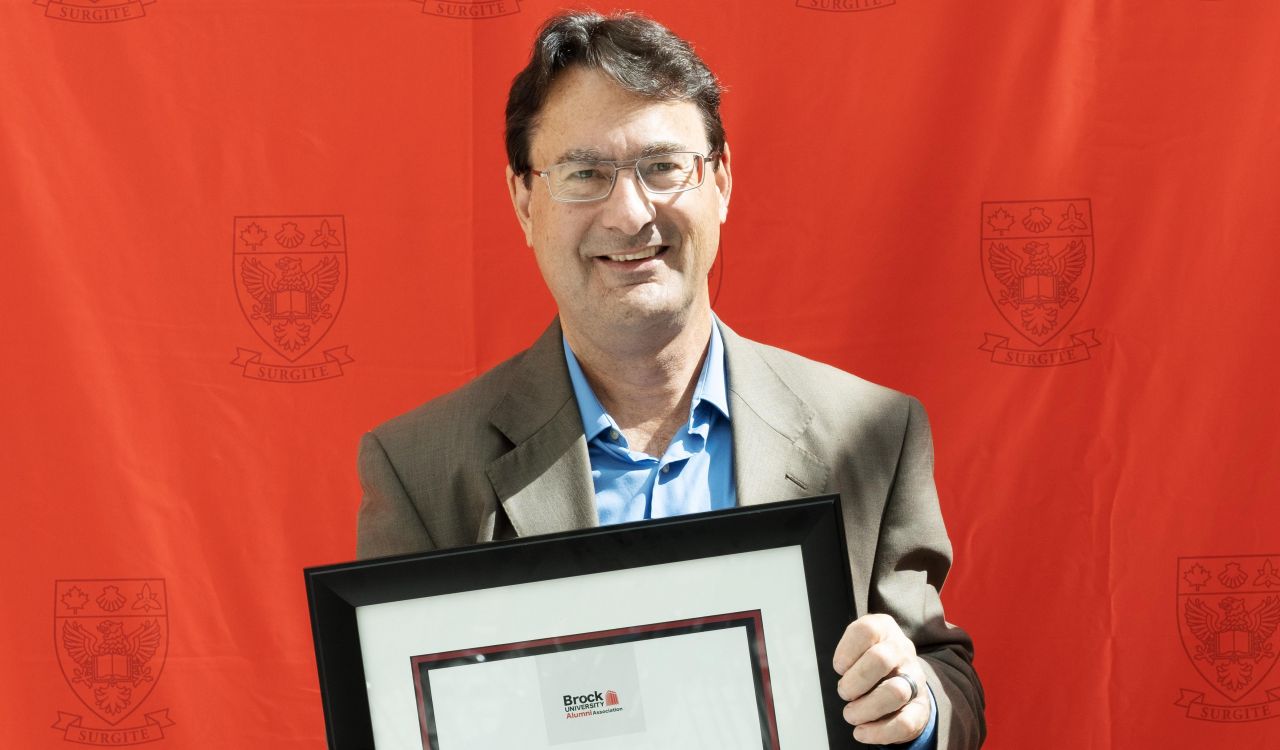 A man holding a framed certificate stands in front of a red backdrop.