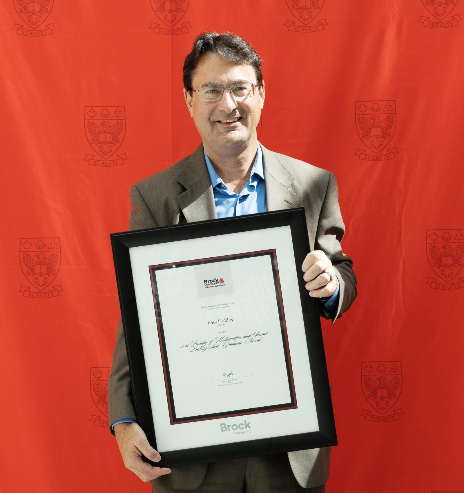 A man holding a framed certificate stands in front of a red backdrop.