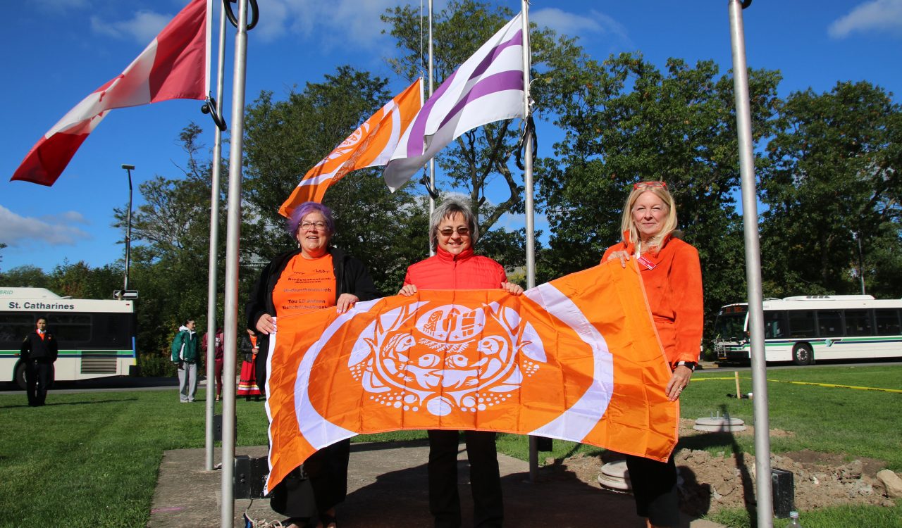 Two women in orange shirts and one in a red shirt stand holding an orange flag with another orange flag and a purle and white flag flying behind them.