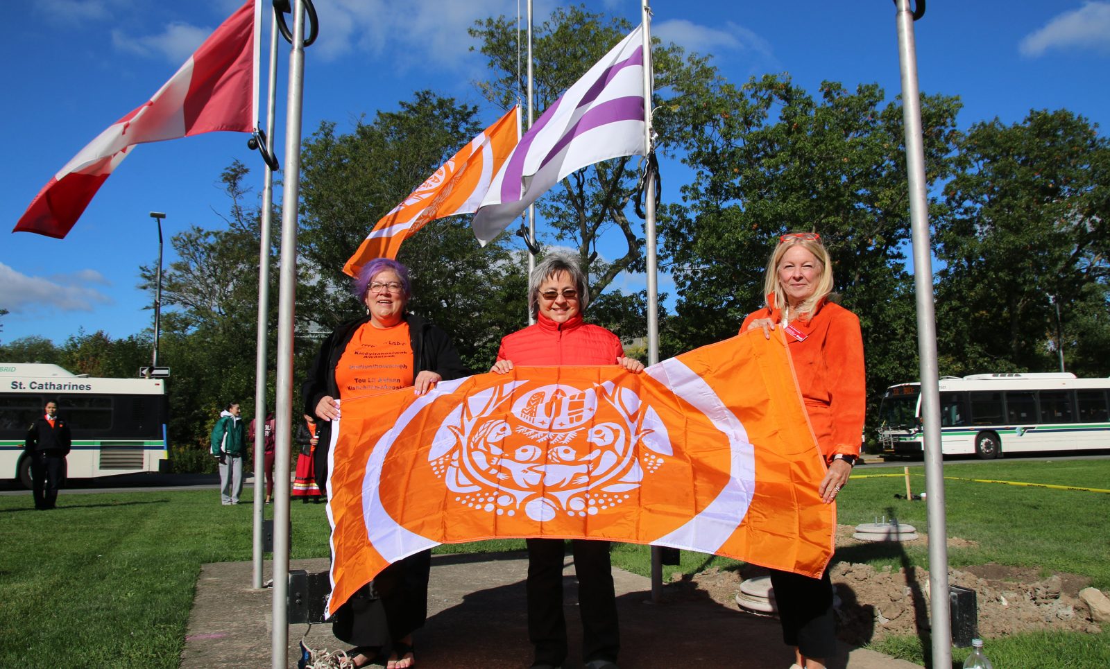 Two women in orange shirts and one in a red shirt stand holding an orange flag with another orange flag and a purle and white flag flying behind them.