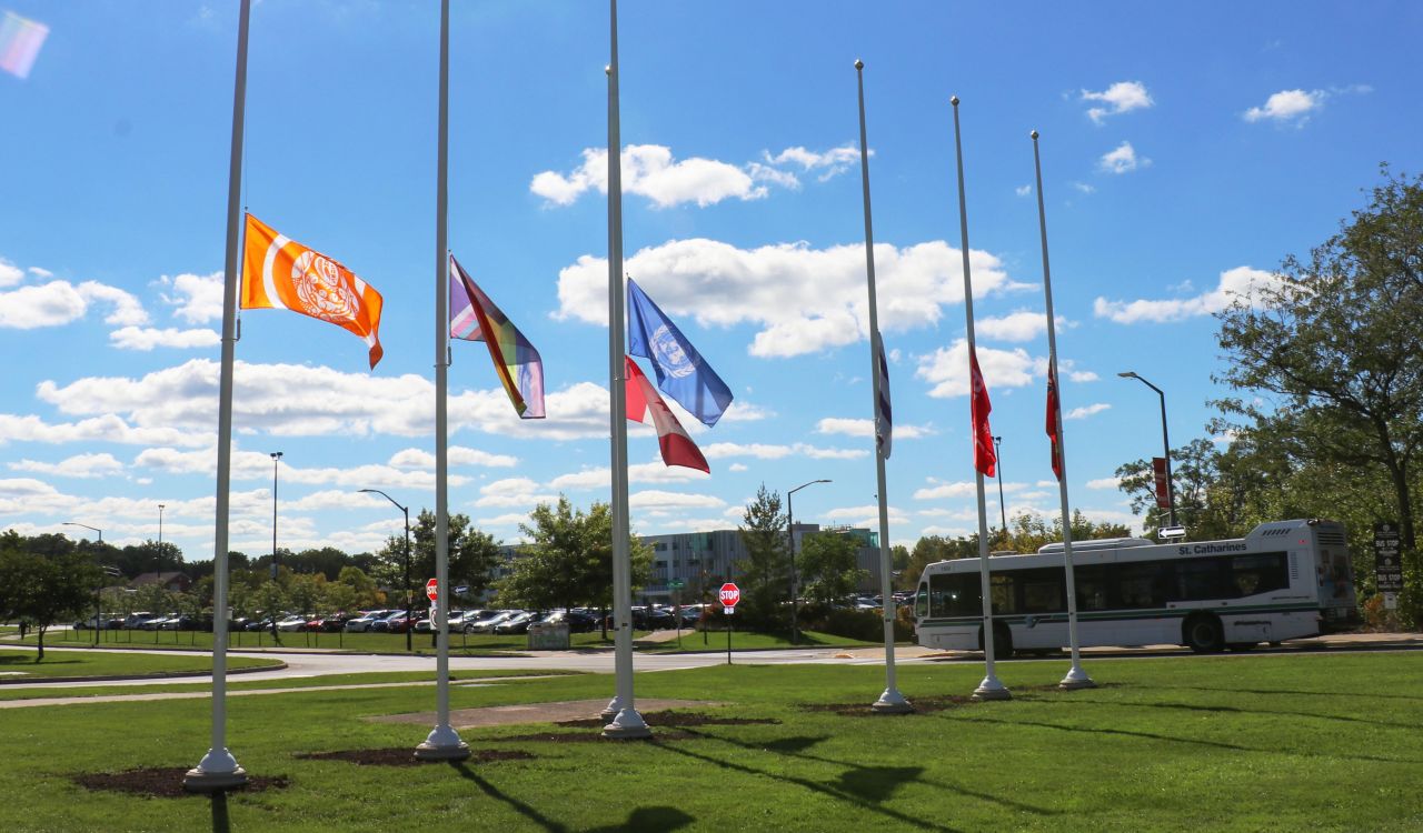 Six flags on separate poles fly at half-mast in front of a blue sky.