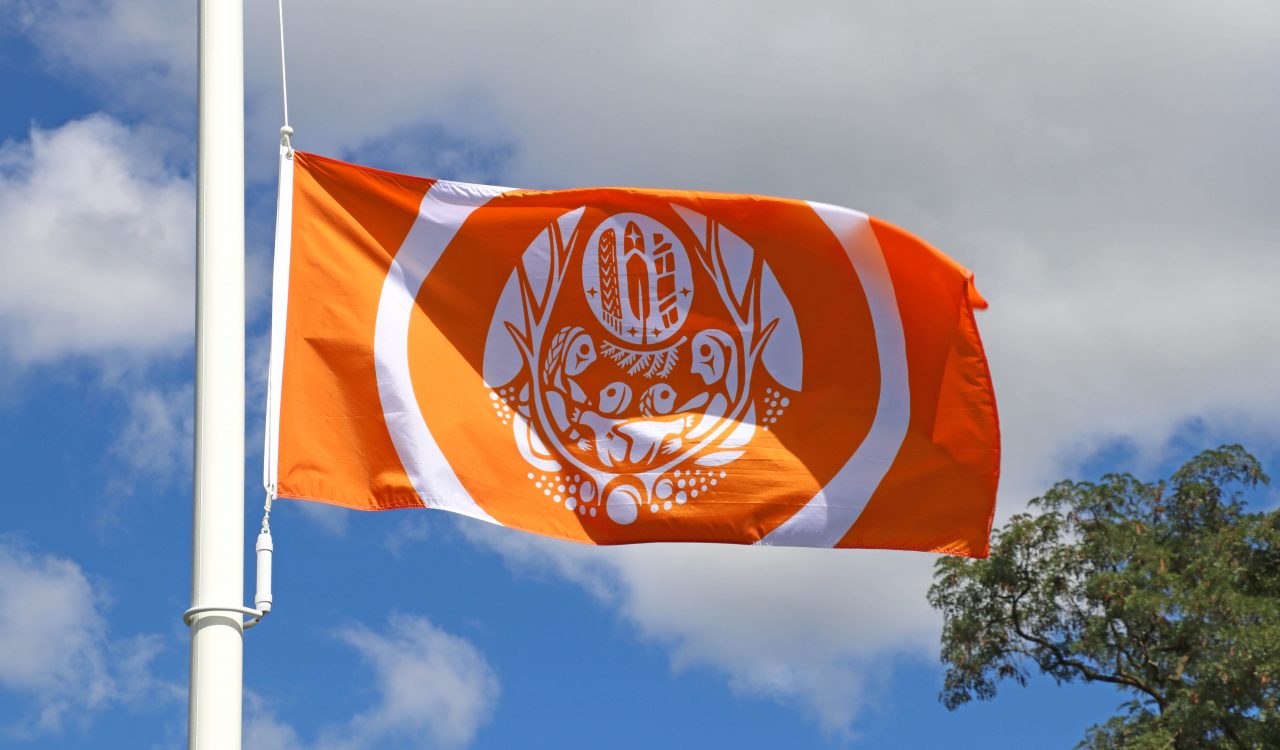 An orange and white flag flies in the wind on a flagpole with trees in the background.