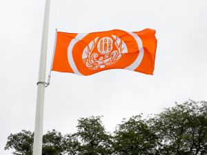 An orange and white flag flies in the wind on a flagpole with trees in the background.