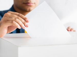 A hand places a ballot into the open section of a ballot box.