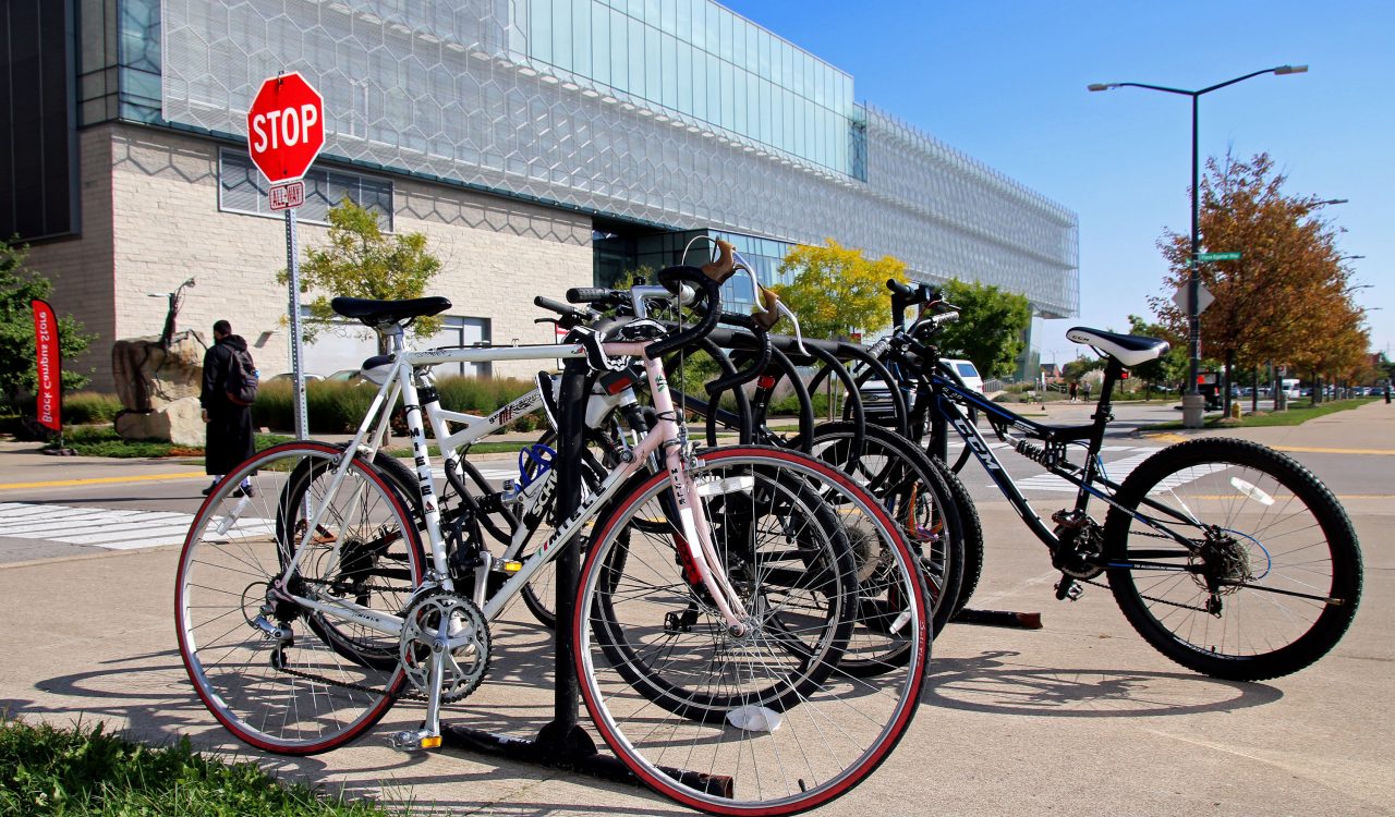 Several bicycles are locked on a tall bicycle rack near Brock University’s Cairns Complex. In the background is a stop sign and a person crossing the street.